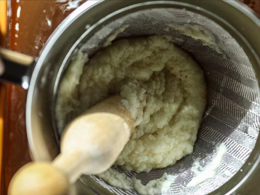 A top-down view of kimchi sauce being pushed through a sieve with a wooden pestle, placed over a metal pot. The mashed potatoes appear creamy and smooth, highlighting the texture achieved through this process.